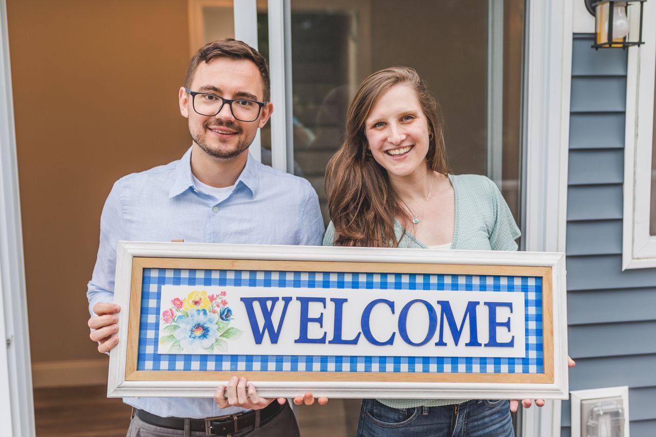 Home A cheerful couple stands at their new home entrance holding a welcome sign.